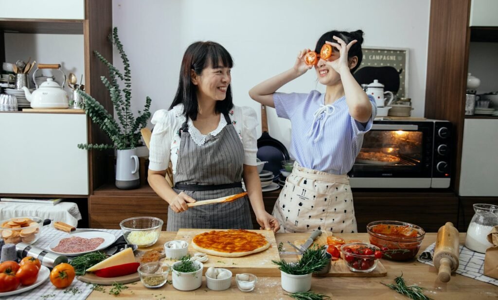 Smiling Asian woman spreading tomato sauce on pizza dough while looking at funny female covering eyes with tomato slices in kitchen